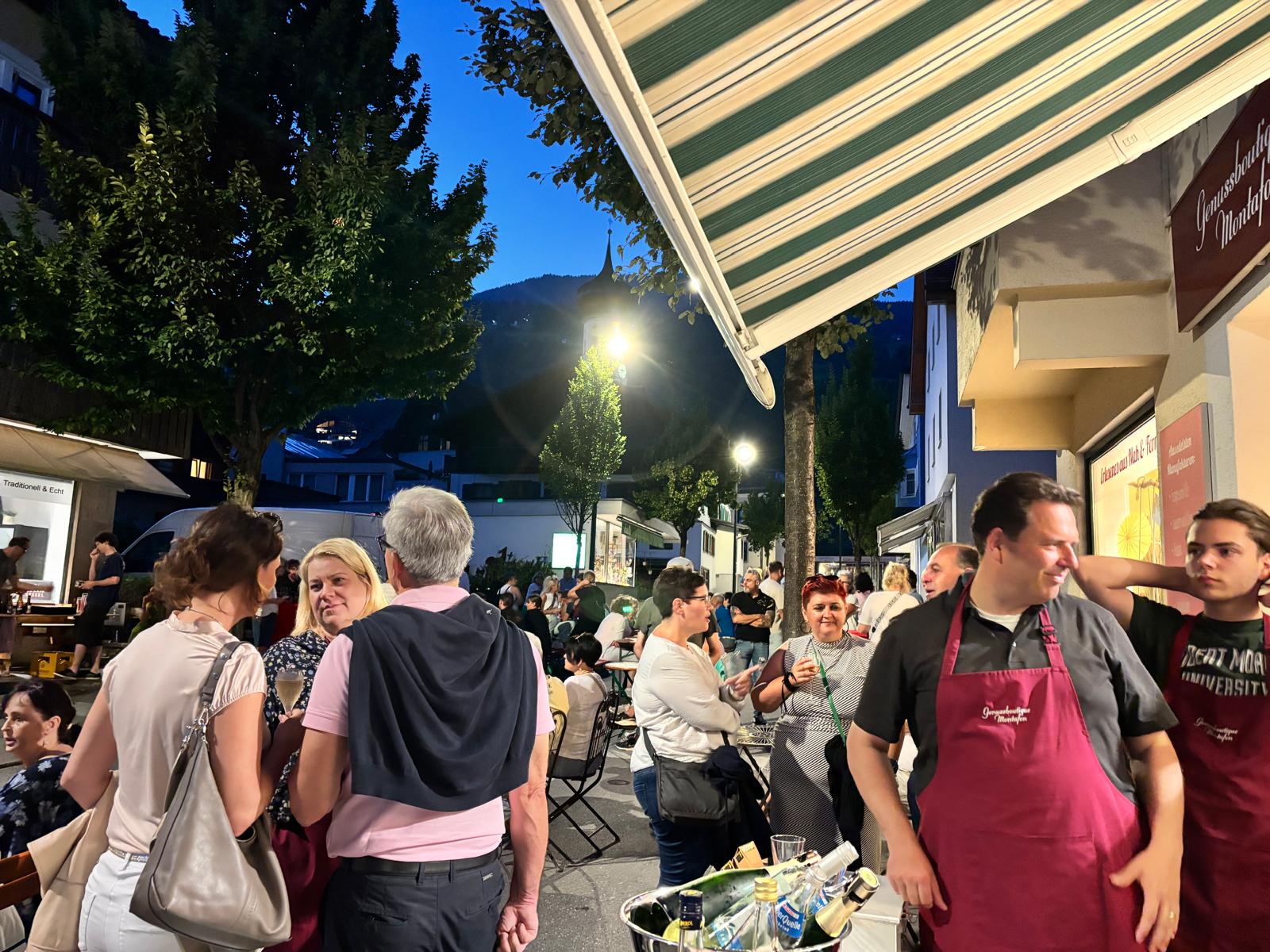 Abendliche Veranstaltung vor der Boutique: Gäste unter dem Vordach, Mitarbeiter in bordeauxroten Schürzen neben einem Eimer mit gekühlten Flaschen