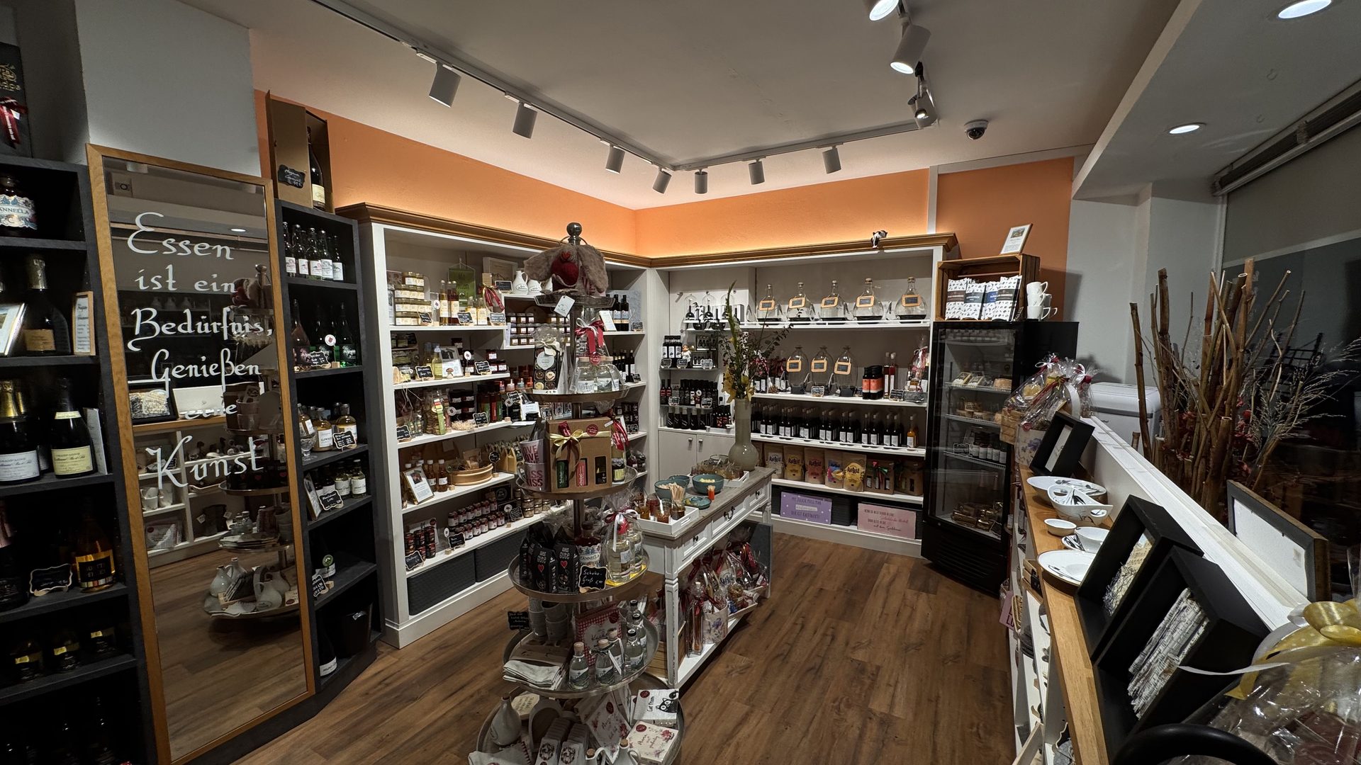Innenansicht der Boutique: Regale und runde Tische mit regionaler Feinkost, Weinen und Geschenkideen, links ein Spiegel mit dem Spruch „Essen ist ein Bedürfnis, Genießen eine Kunst“